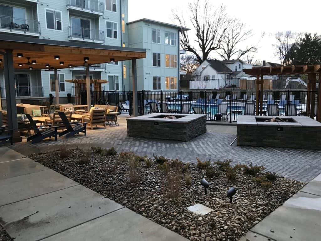 Elegant outdoor living space featuring a covered seating area, stone fire pits, and surrounding landscaping, designed for relaxation and entertaining, with a view of a pool in the background.