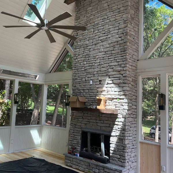 Custom stone fireplace with wooden mantel in a bright, open sunroom, featuring large windows and ceiling fan, enhancing outdoor living space ambiance.