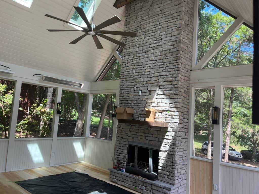 Sunroom interior featuring a stone fireplace, large windows with natural light, and a ceiling fan, showcasing a cozy outdoor living space designed by DW Construction.