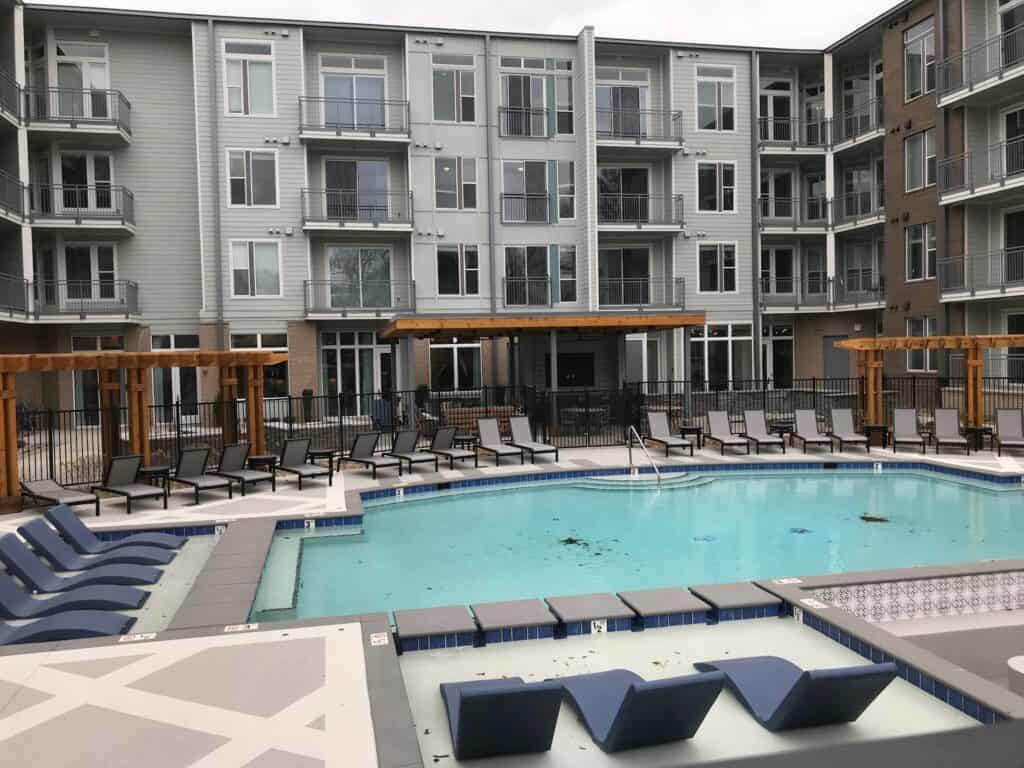Luxury apartment complex featuring a spacious outdoor pool area with lounge chairs, surrounded by modern architecture and a wooden pergola, emphasizing upscale outdoor living spaces.