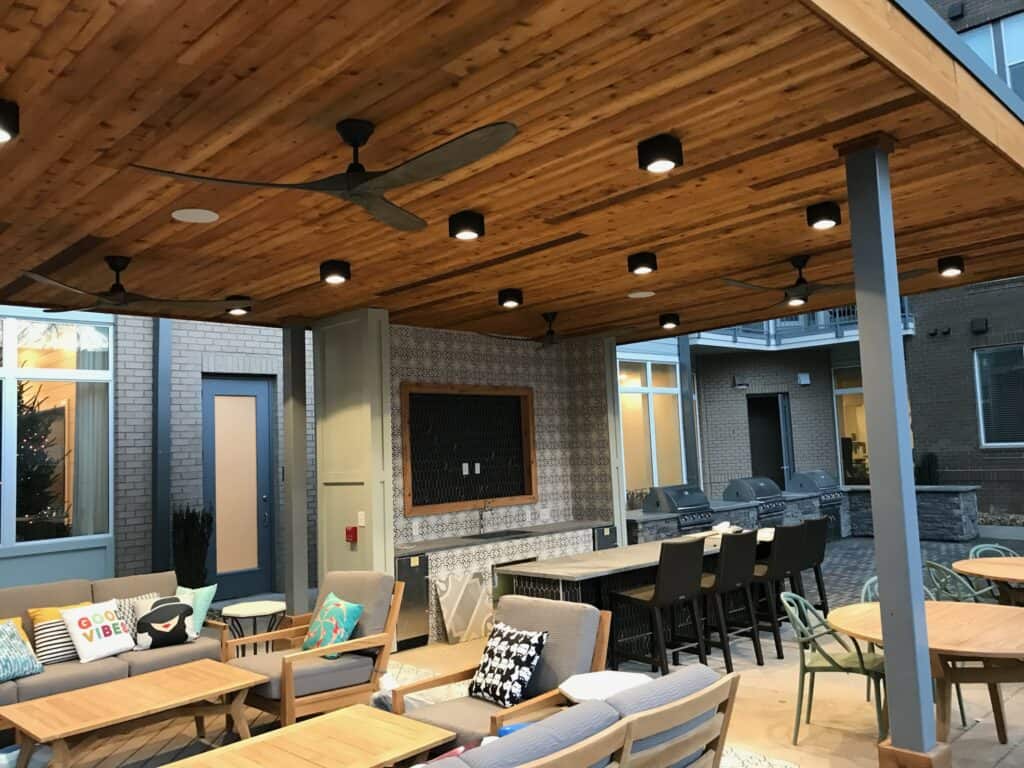 Outdoor living space featuring a wooden ceiling with lights and fans, comfortable seating with decorative pillows, dining area with tables and chairs, and a backdrop of modern architecture, showcasing DW Construction's design expertise in patios and porches.