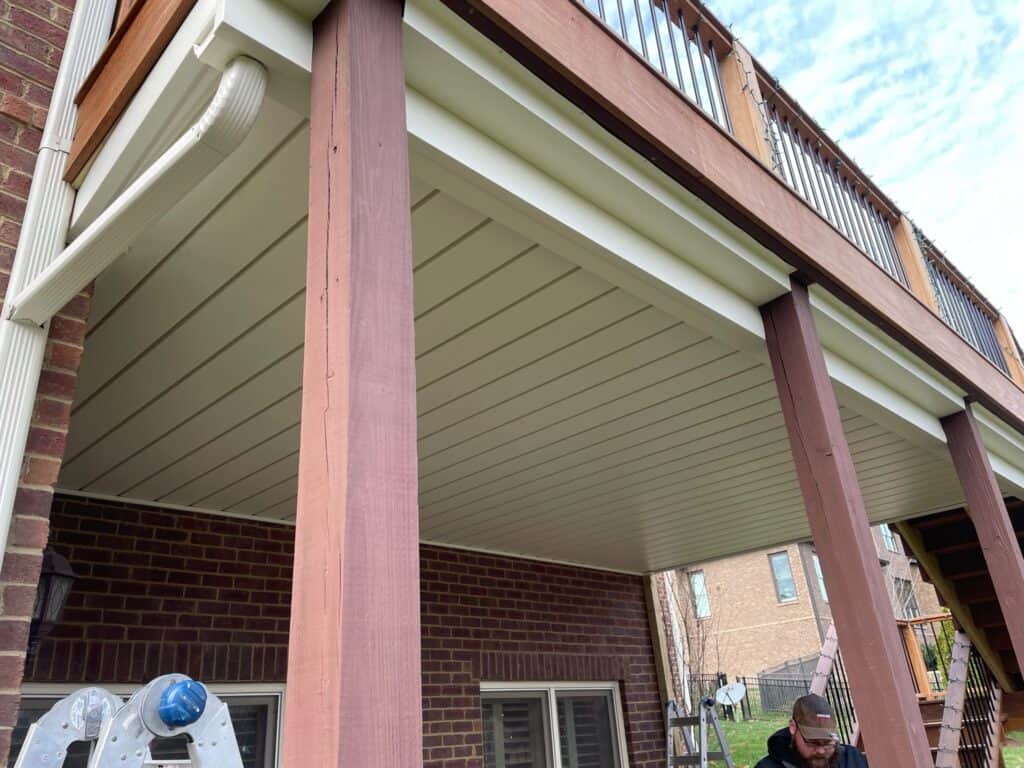 Underneath a wooden deck featuring vertical support beams and a finished ceiling, with a contractor working on-site in a residential backyard setting.