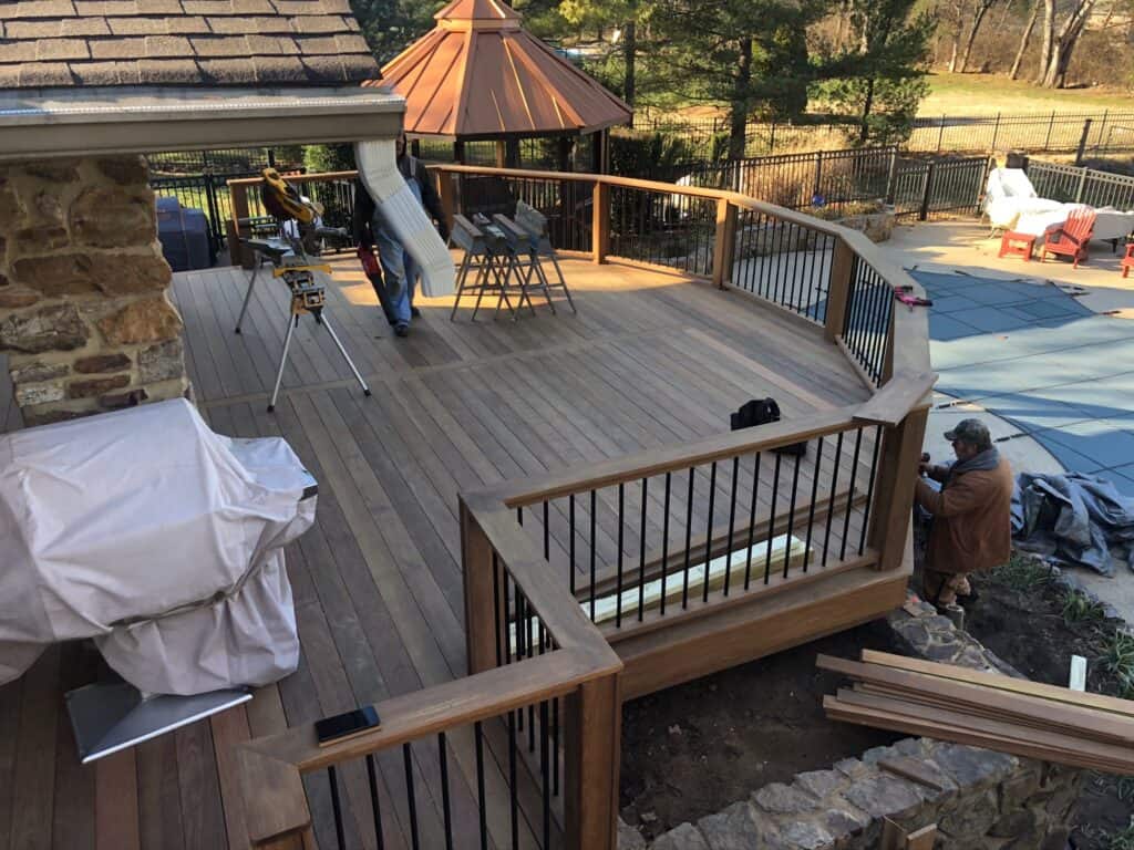 Custom wooden deck under construction with workers, featuring a stone wall, black railings, and tools, showcasing DW Construction's outdoor living project expertise.