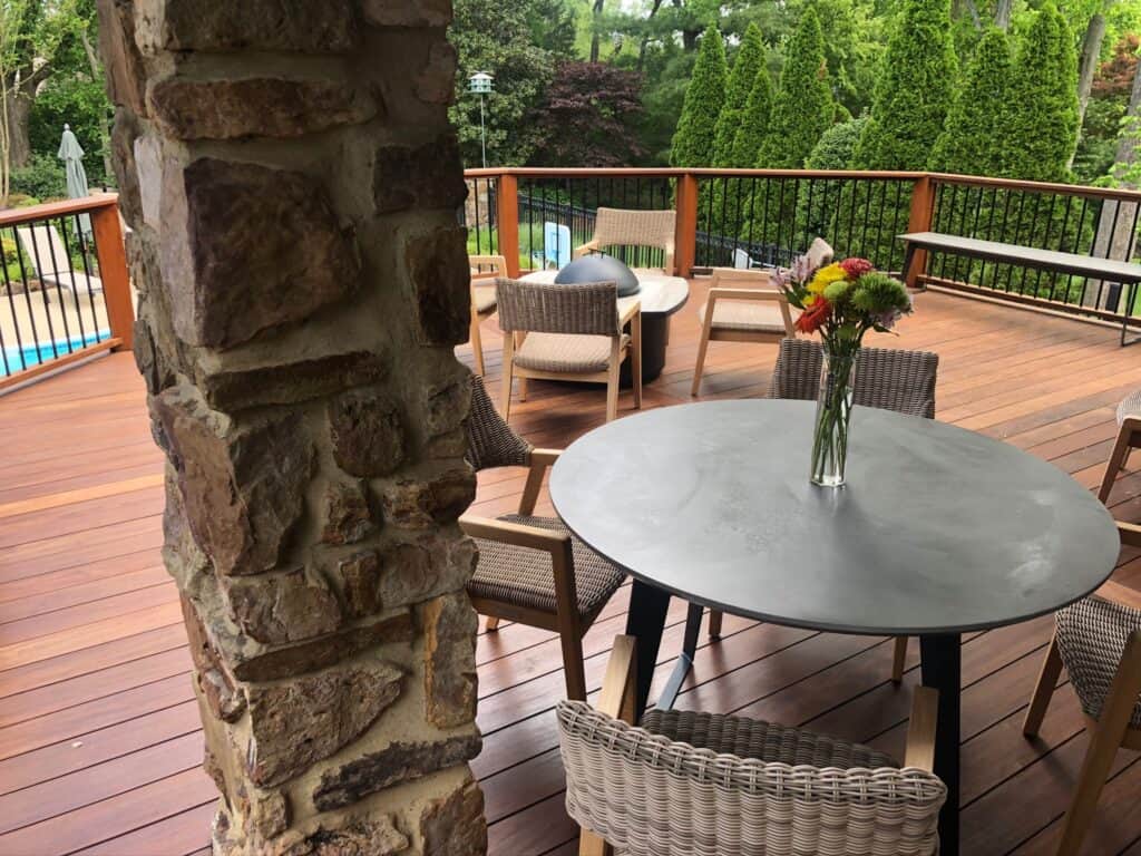 Outdoor living space featuring a stone pillar, round table with flowers, and wicker chairs on a wooden deck, surrounded by greenery and a pool, showcasing DW Construction's custom patio design.
