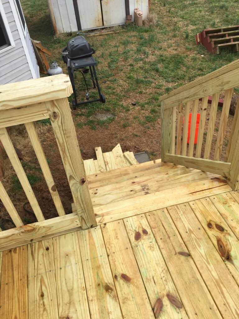 Newly constructed wooden deck with stairs leading to a grassy area, featuring railings, a grill, and a shed in the background.