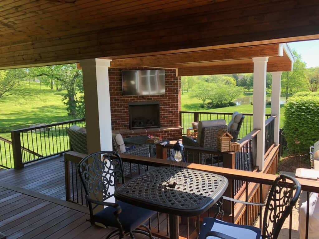 Outdoor living space featuring a covered deck with a dining table, comfortable seating area, a brick fireplace, and a mounted TV, surrounded by lush greenery in Nashville, TN.