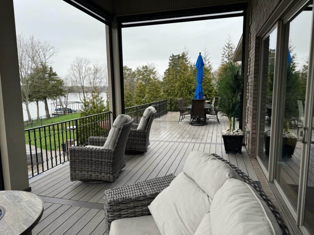 Outdoor living space featuring a deck with wicker seating, a dining area under a blue umbrella, and views of a lake surrounded by trees, showcasing DW Construction's custom design and build capabilities.