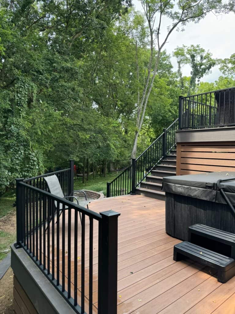 Outdoor wooden deck with black railings, hot tub, and lounge chair, surrounded by lush greenery, showcasing DW Construction's custom outdoor living design.