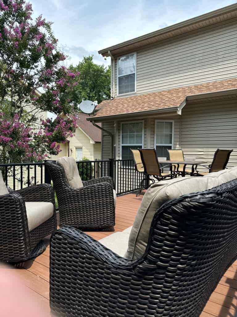 Outdoor living space featuring comfortable wicker chairs with cushions on a wooden deck, surrounded by flowering trees and a view of a home, showcasing a relaxing setting for entertaining or leisure.